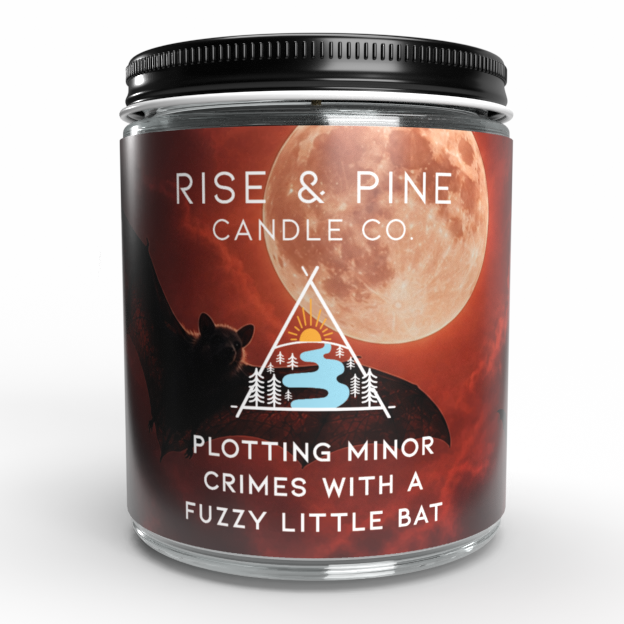A glass jar candle with a black metal lid and a dark red label featuring a large full moon, a black silhouette, and the brand name 'RISE & PINE CANDLE CO.' The label also reads 'PLOTTING MINOR CRIMES WITH A FUZZY LITTLE BAT.